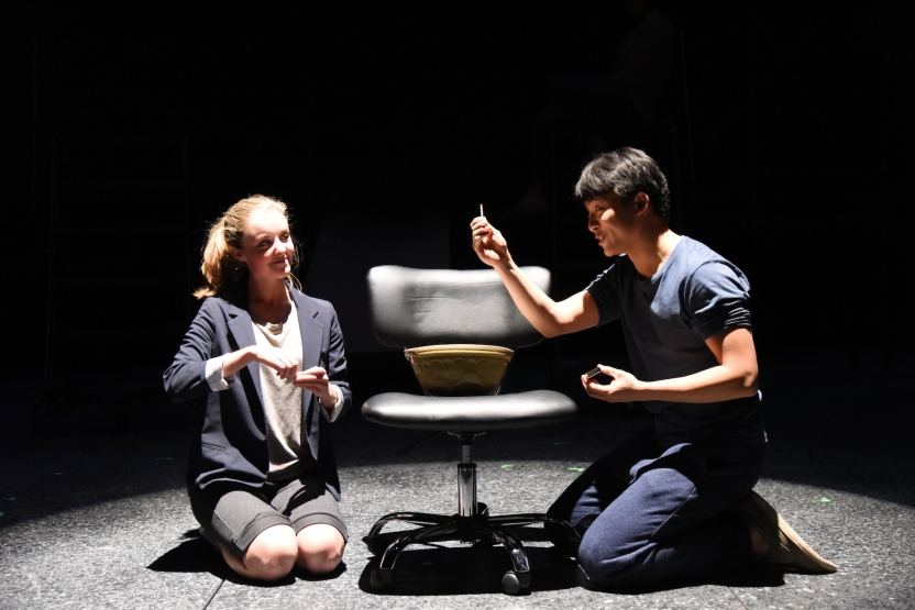 Two actors kneeling on each side of a rolling chair, one with a match outstretched