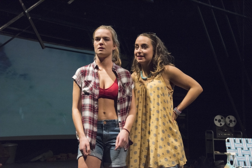 Two actresses standing side by side, one with yellow dress, another with flannel and red bra
