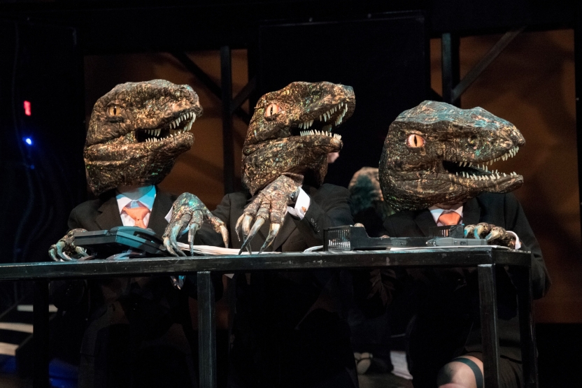 Three actors wearing dinosaur heads at a desk facing right