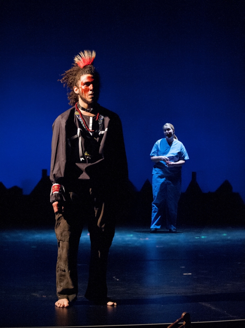 Actor with red face paint and headpiece at forefront, nurse in background