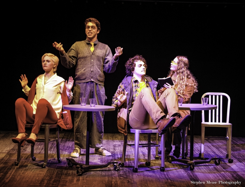 Two actors in chairs with hands and feet up like dogs, one actor standing between them talking as another takes notes