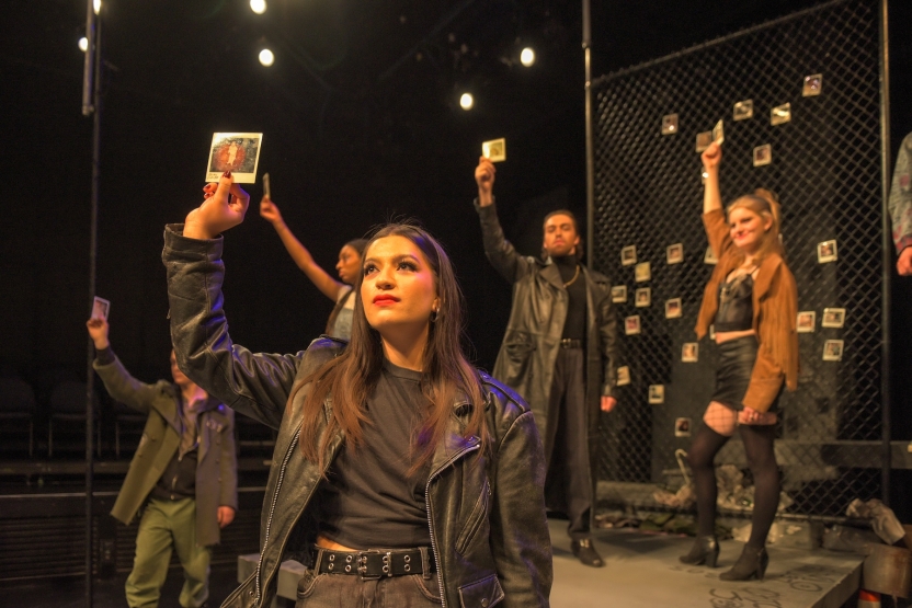People wearing mostly black and leather scattered across the stage and holding up polaroids
