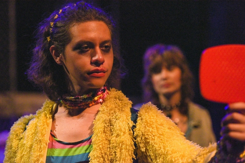 Person looking in the mirror with smudged makeup as another person looks at them in the background