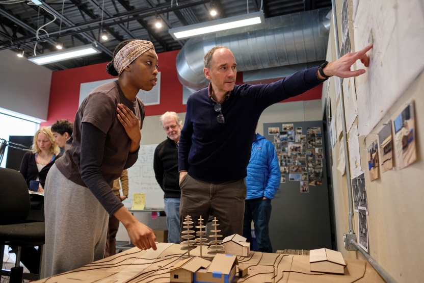 A student and professor look over a cardboard model. The professor points something out on a whiteboard as the student looks on.