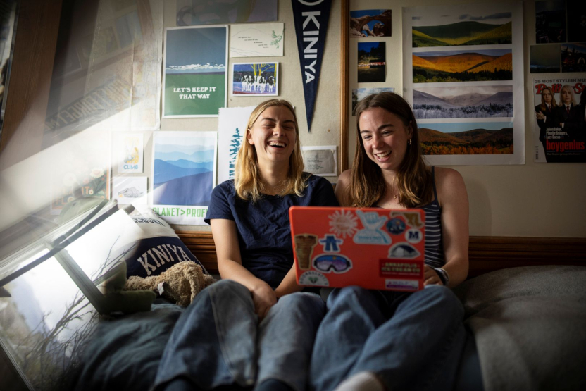 Two students look at a laptop and laugh as they sit on a bed in a dorm room. 