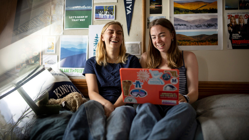 Two students look at a laptop and laugh as they sit on a bed in a dorm room. 