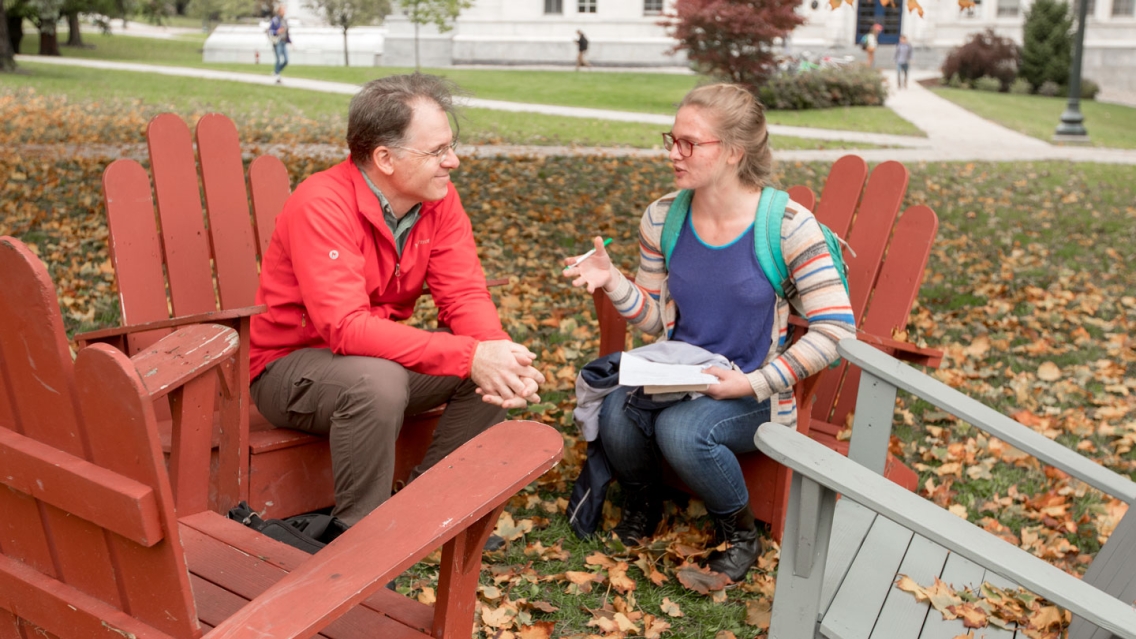 A professor and student sit in chairs outside on a fall day to discuss a project.