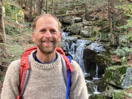 Photo of Professor Marc Lapin in the woods in front of a waterfall with a backpack on.