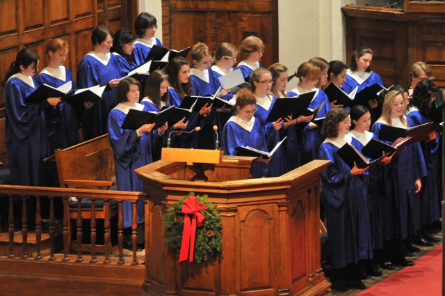 A group of choir singers in blue robes, in Mead Chapel, with a holiday wreath