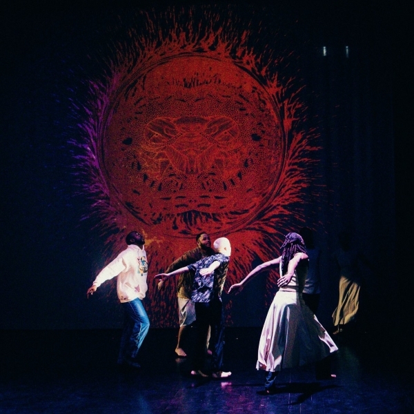 Three dancers on a dark stage with a red artifact image projected behind them (171)
