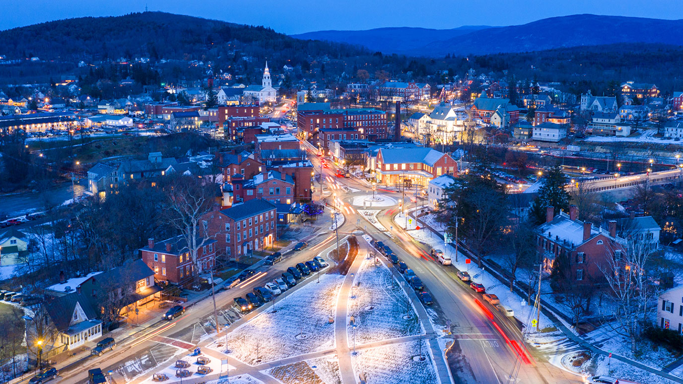 An aerial photo of downtown Middlbury, Vermont in the winter at night. Lights can be seen from cars and red brick buildings which line the street. In the foreground is a park with a light covering of snow.