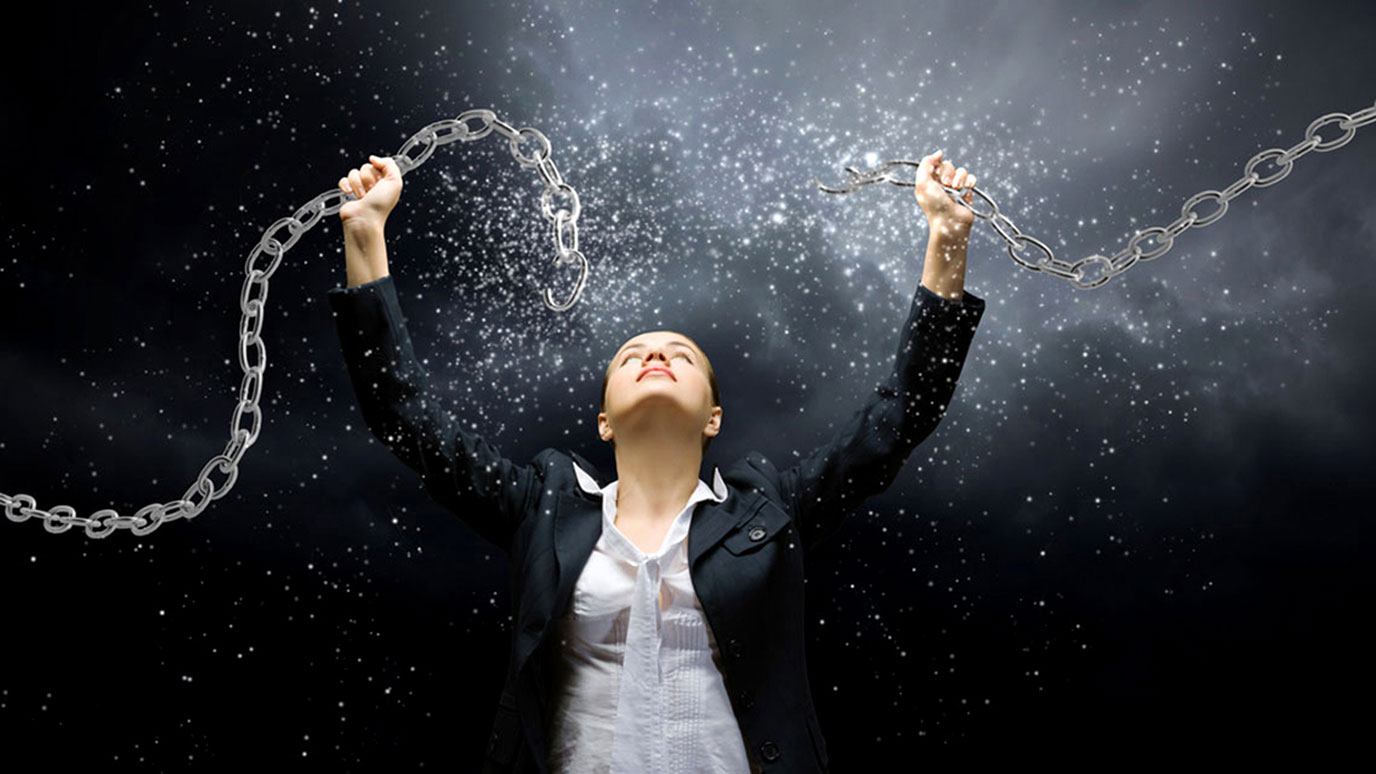 A woman wearing a suit stands looking up at two segments of a chain she is holding above her head. She appears to have broken the chain and sparks and debris are flying out of it.