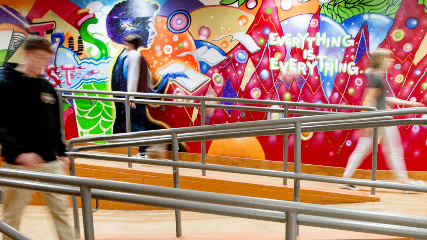 People are seen walking down a ramp in front of a colorful mural which contains the words 'Everything is Everything'. The people are blurry, indicating movement.