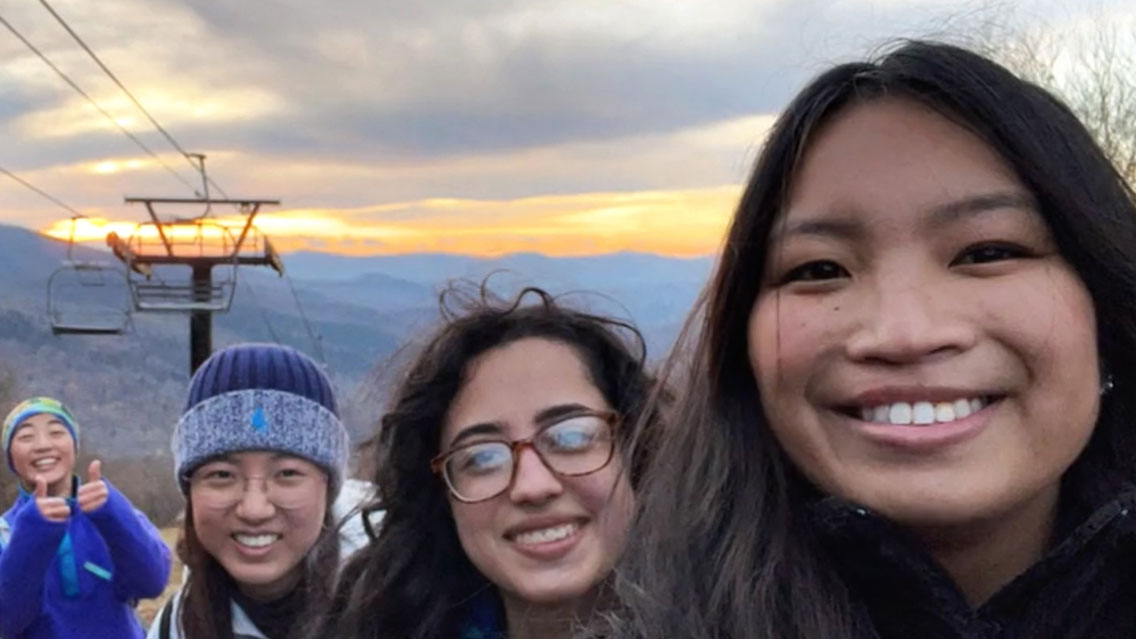 The heads of four women are seen, smiling while they stand in front of a chairlift at a ski slope.