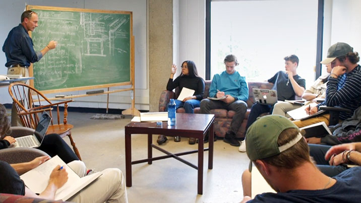 A man stands at a chalkboard covered in diagrams in front of a room of college students sitting on couches in a horseshoe layout taking notes.
