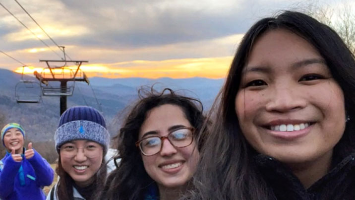 The heads of four women are seen, smiling while they stand in front of a chairlift at a ski slope.