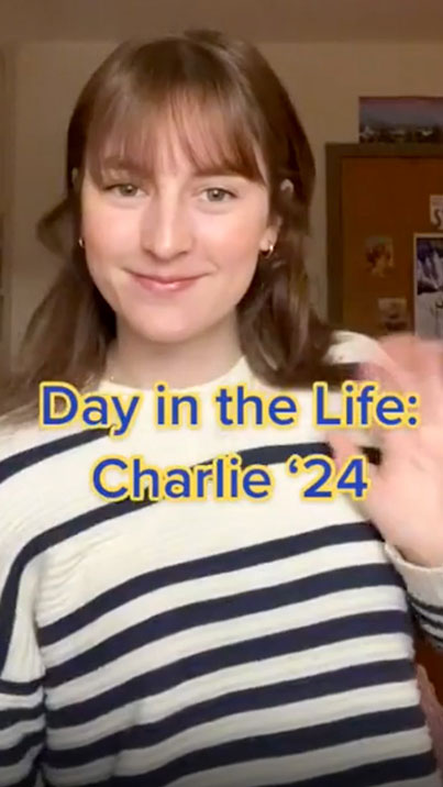 A portrait photo of a woman wearing a white shirt with black horizontal stripes smiling with her left hand raised waving toward the camera.