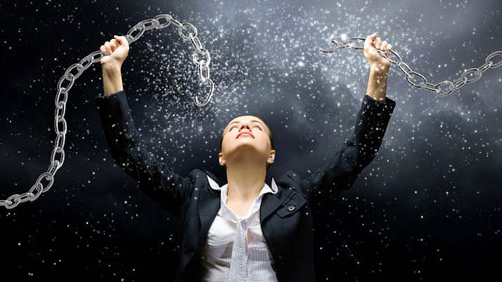 A woman wearing a suit stands looking up at two segments of a chain she is holding above her head. She appears to have broken the chain and sparks and debris are flying out of it.
