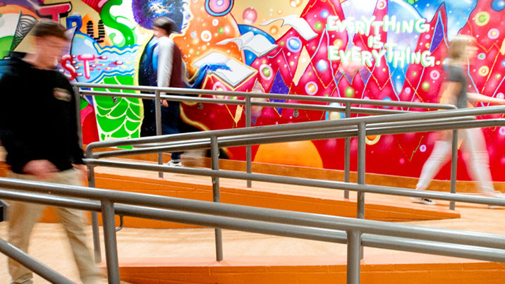 People are seen walking down a ramp in front of a colorful mural which contains the words 'Everything is Everything'. The people are blurry, indicating movement.