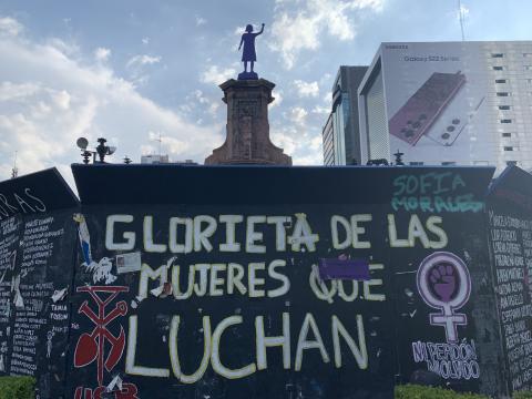 Photograph of a black wall with white painted letters reading "Glorieta de las mujeres que luchan." Statue of a person raising a fist can be seen above the wall against a blue sky with clouds.