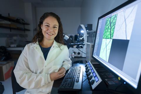 Woman in white lab jacket standing in from of large computer screen.