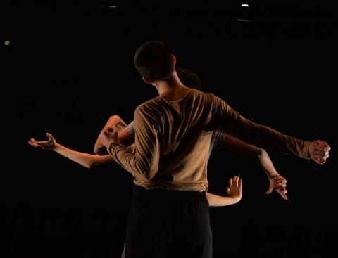 Three dancers standing in a line, as seen from behind, against a dark background