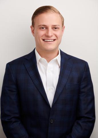 A man with short blond hair in a blue suit jacket and white dress shirt, smiling.