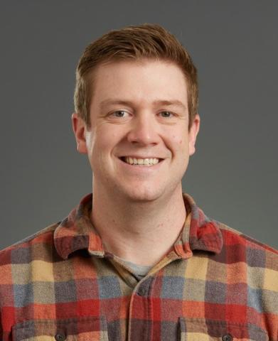 Man smiling wearing orange, gold, grey plaid shirt.