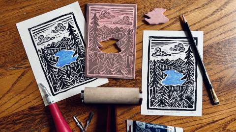 Linocut stamp and prints arranged on wooden table with ink roller and linocut materials scattered around.