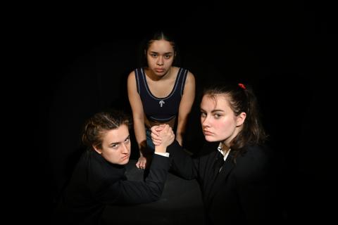 Three woman, two of them clasping hands as in an arm-wrestling match, with black background