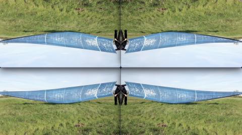 A digitally manipulated image divided into four mirrored quadrants. The top half of the image is an upside-down mirror of the bottom half, and the left side mirrors the right, creating a symmetrical, kaleidoscope-like effect. The primary image shows a person dressed in black, standing on green grass, holding the edge of a large, translucent blue piece of fabric with white markings that stretches out horizontally.