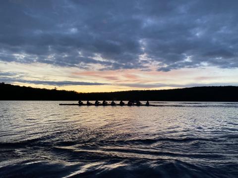 An 8-shell of rowers cuts across a serene Lake Dunmore with the setting sun visible in the background.