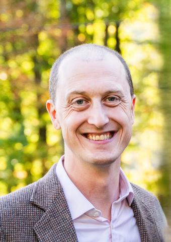 Man in pink shirt, brown jacket smiling. Trees in background.