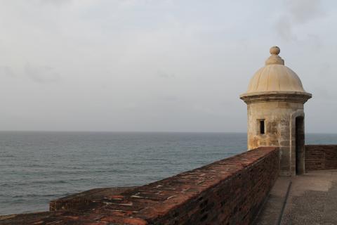 A part of the wall and a white stone turret of the fort in Old San Juan stands in the foreground with the Atlantic Ocean behind it.
