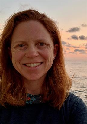 Woman with red, should length hair. Blue sweater, ocean in background.