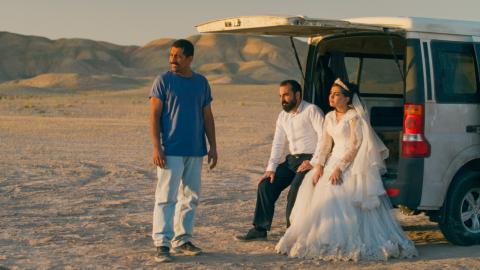 Three people are watching a sunset in the middle of a desert. Two of them sit in the open trunk of a truck and are dressed as the groom and the bride.