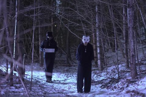 Two people in snowy woods, one wearing a mask and facing the camera, one facing away from the camera