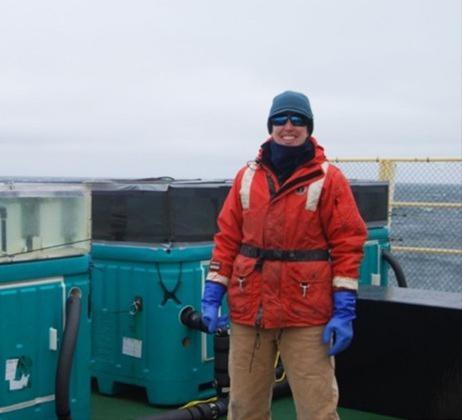 Image of Casey Schine in orange coat, hat, gloves, and sunglasses on a boat with the ocean and cloudy sky behind her.