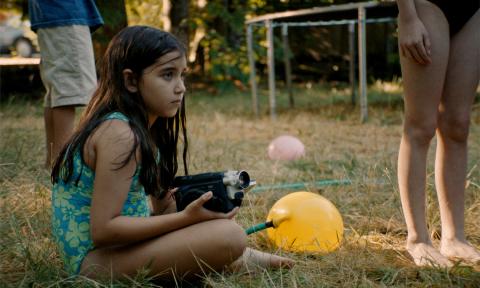 A young girl in a swimsuit crouches on dry grass, holding a video camera, looking off to the side. Nearby are other children’s legs, a yellow water balloon attached to a hose, getting filled with water.