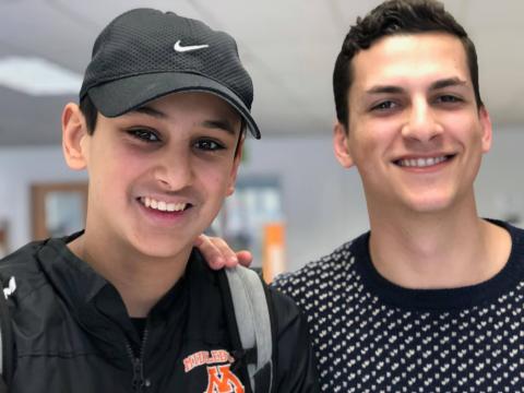 Two people stand next to each other, one with their hand on the other's shoulder. They are both smiling at the camera.