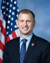 Image of a man wearing a blue tie standing in front of an American flag