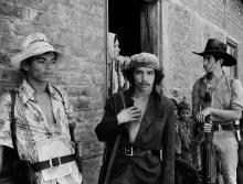 A black and white photograph of three Salvadoran men standing in front of a stone building, each looking in a different direction.