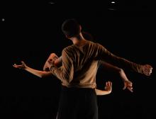 Three dancers standing in a line, as seen from behind, against a dark background