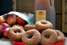 red plate of cider donuts with cinnamon sugar, tipped open bag of apples and 1/2 gallon plastic jug of cider in background. Wood wall behind.