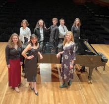 students on stage standing around a piano