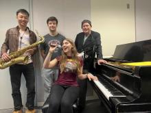 Four people standing near a piano; one is holding a saxophone