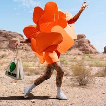 Person dressed in an orange costume consisting of upward facing panels that obscure the upper body and head, seemingly in motion walking through an arid landscape with one arm raised and with the other holding a canvas bag.