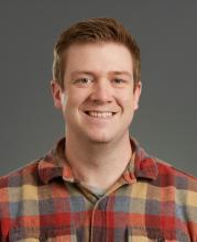 Man smiling wearing orange, gold, grey plaid shirt.