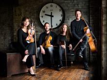 Members of the quartet seated, holding their instruments, in front of a large clock