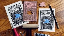 Linocut stamp and prints arranged on wooden table with ink roller and linocut materials scattered around.